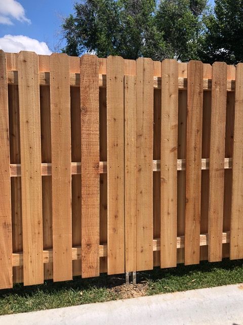 A new, light brown wooden privacy fence with vertical pickets, set against a background of trees and a blue sky.