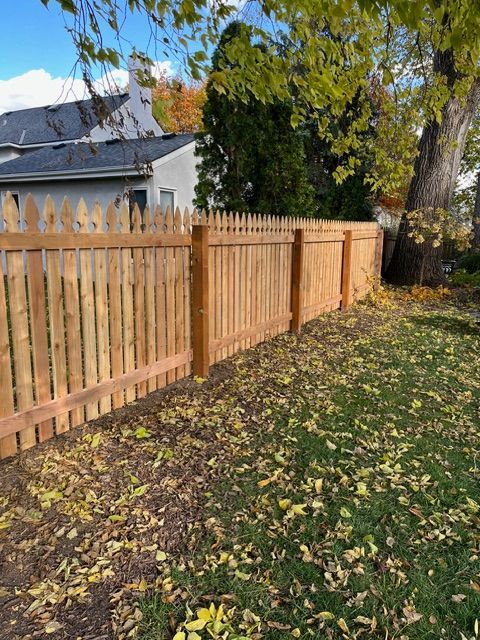 A newly installed wooden picket fence stands in a backyard scattered with fallen autumn leaves next to a house.