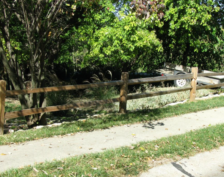A wooden split-rail fence runs along a concrete sidewalk next to green trees and foliage in a sunny outdoor setting.