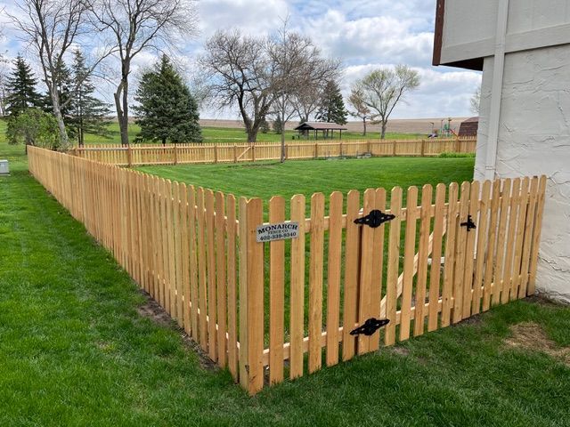 A newly installed wooden picket fence with a closed gate, bordering a green lawn in a rural outdoor setting.