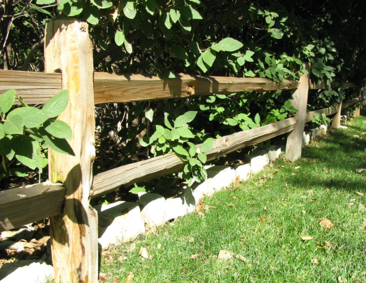 A rustic wooden split-rail fence runs along a green grassy lawn with light-colored stones lining the base.