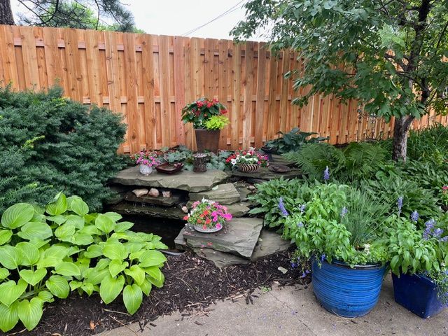 A tiered stone garden water feature sits in front of a wooden fence, surrounded by lush hostas and potted flowers.