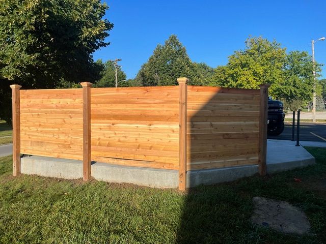 A wooden horizontal slat privacy fence built on a concrete curb, surrounded by grass under a clear blue sky.