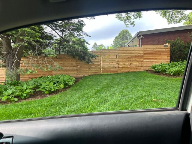 A wooden horizontal-slat fence runs across a grassy backyard with plants, viewed from inside a car window.