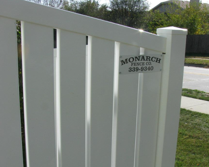A white vinyl fence gate with a Monarch Fence Co. plaque attached to the corner post, seen from an outdoor sidewalk.
