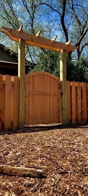 A wooden arched gate integrated into a fence, topped with a decorative wooden pergola structure in a yard with mulch.