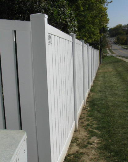 A long, white vinyl privacy fence lines the side of a grassy residential lawn next to a paved road.