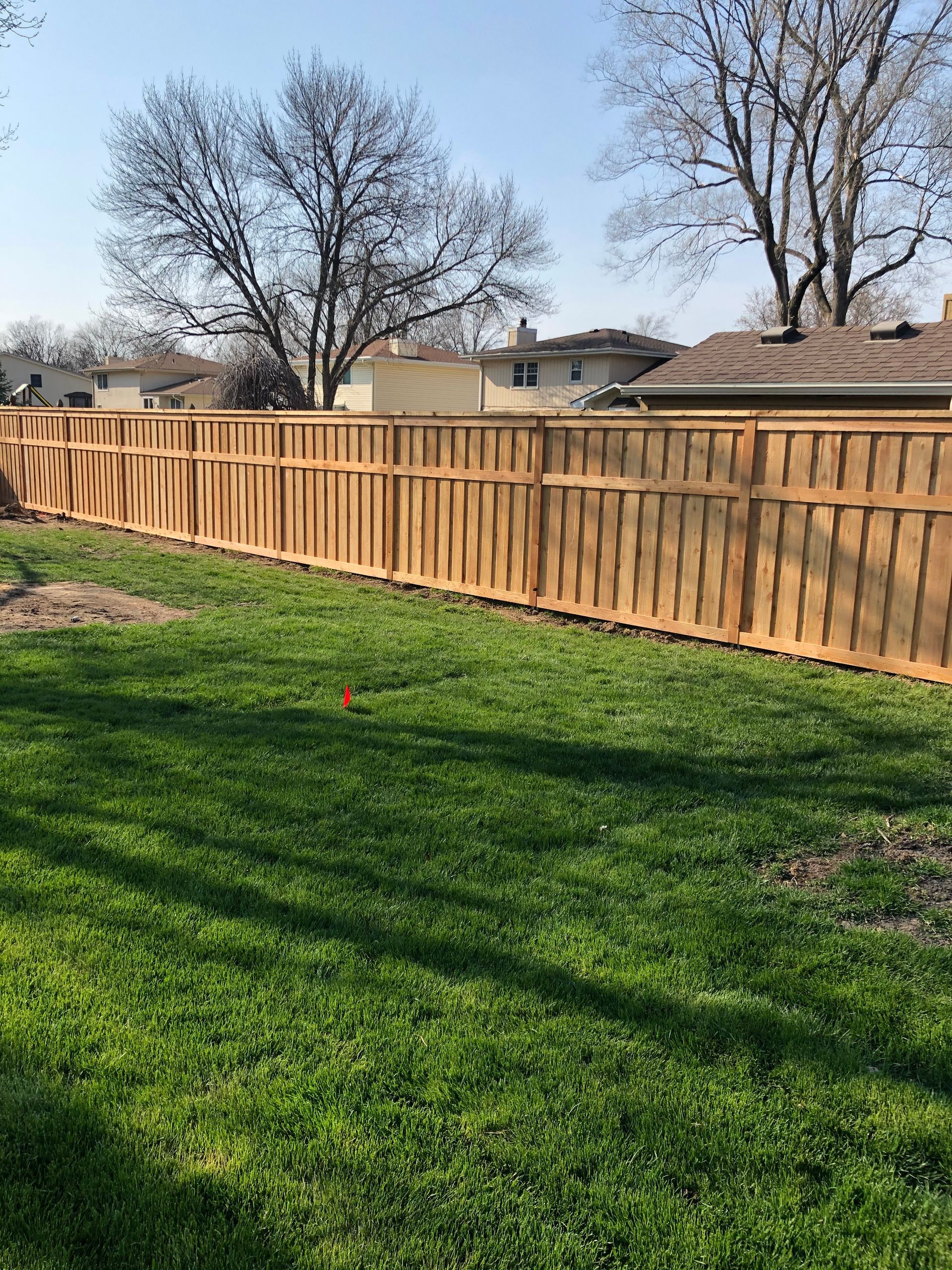 A wooden fence lines the back of a green, grassy yard on a sunny day with trees and neighboring houses in the background.