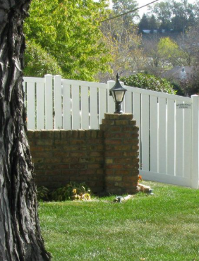 A short, brick wall with a topped lamp post and white vinyl fencing, situated in a green, grassy yard next to a tree.