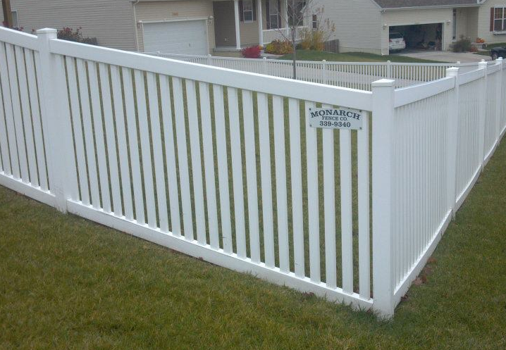 A white vinyl fence with a contractor’s sign on a post, situated on a green lawn in front of suburban houses.