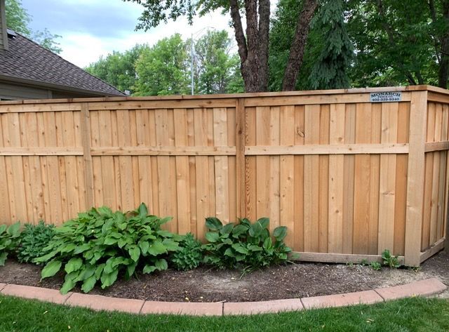 A wooden privacy fence borders a garden bed with hostas and a curved, concrete paver edge in a backyard.