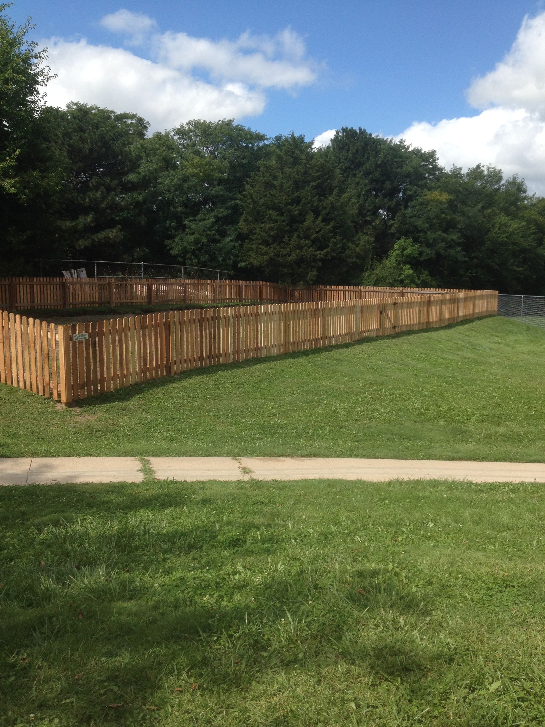 A wooden picket fence encloses a green grassy area on a sunny day with trees in the background.
