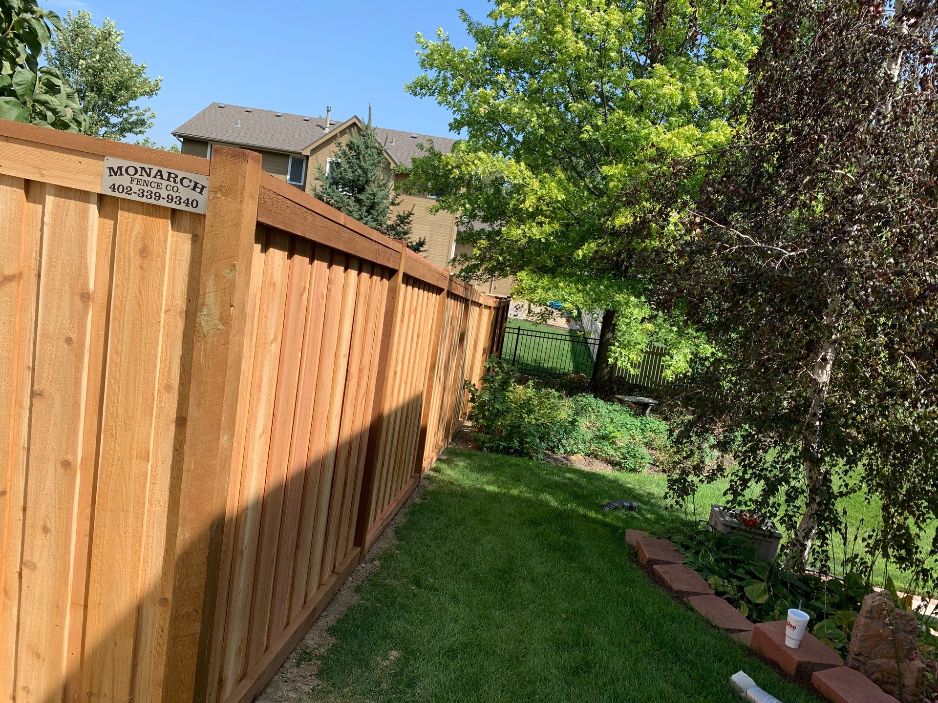 A wooden privacy fence borders a grassy backyard on a sunny day with trees and a neighboring house in the background.