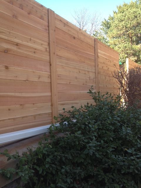 A tall, horizontal-slat cedar wood fence with vertical posts, bordered by a green bush and a white ground-level trim.
