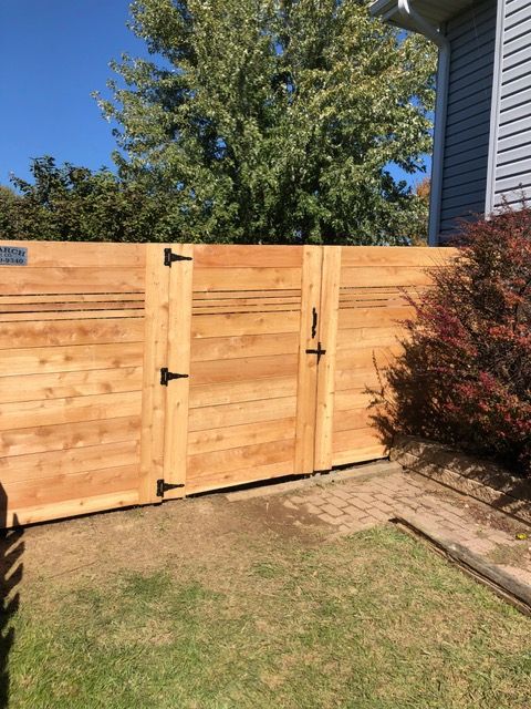 A wooden gate with black hinges and a latch, set in a fence next to a house and a bush on a sunny day.