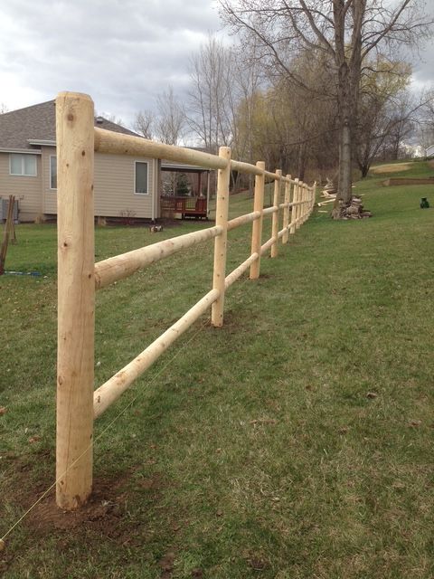 A new wooden three-rail post-and-rail fence stands on a grassy lawn in front of a residential house.