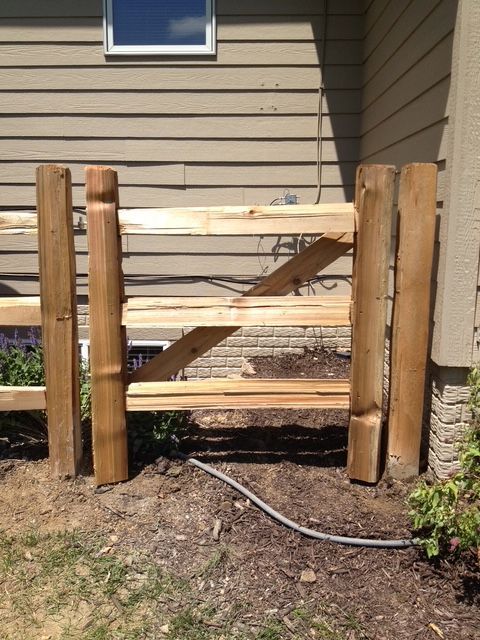A wooden gate made of horizontal rails and vertical posts sits in front of a house with tan siding and a brick foundation.