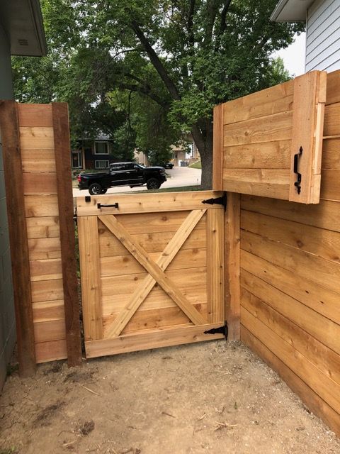 A wooden gate with an X-brace design installed in a fence, featuring black hinges and a latch against a gravel ground.