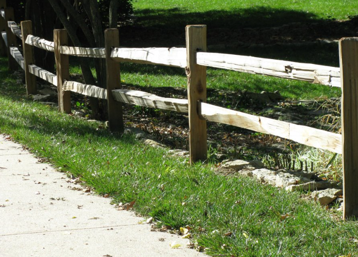 A wooden two-rail fence runs along a concrete sidewalk beside a grassy lawn.