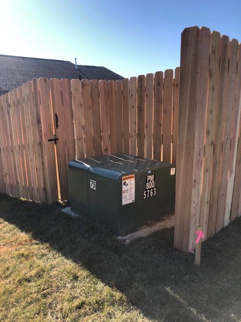 A green pad-mounted electrical transformer sits on a concrete slab in a yard, partially enclosed by a wooden fence.