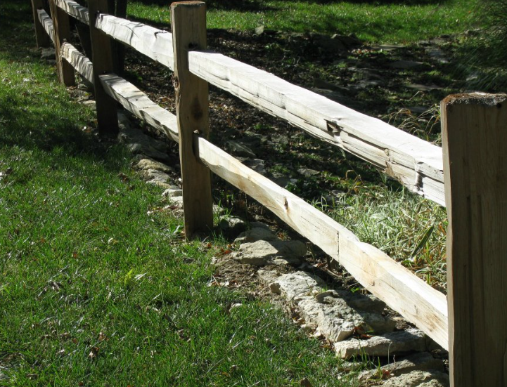 A wooden post-and-rail fence runs diagonally through a grassy yard with a stone border at its base.