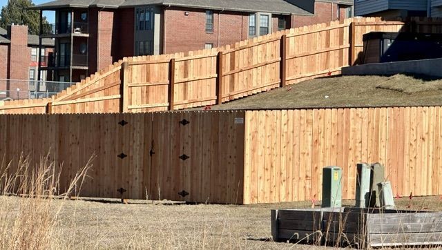A new light-wood privacy fence borders a grassy hill in front of a red brick apartment building.