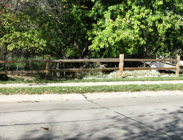 A two-rail wooden fence lines a sidewalk next to a road, with dense green trees in the background.