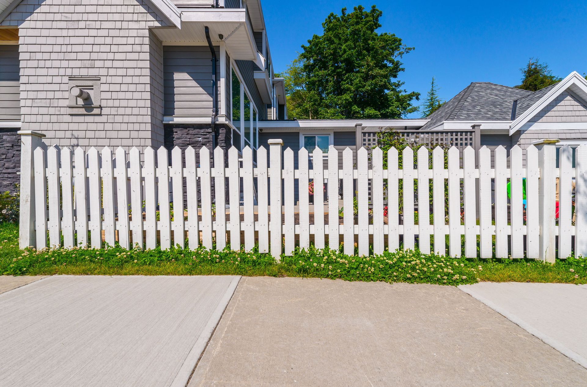 A white picket fence stands in front of a gray house with a gray shingled roof on a bright, sunny day.