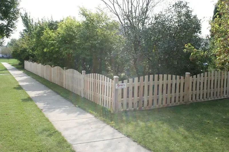 A wooden picket fence along a sidewalk with trees in the background