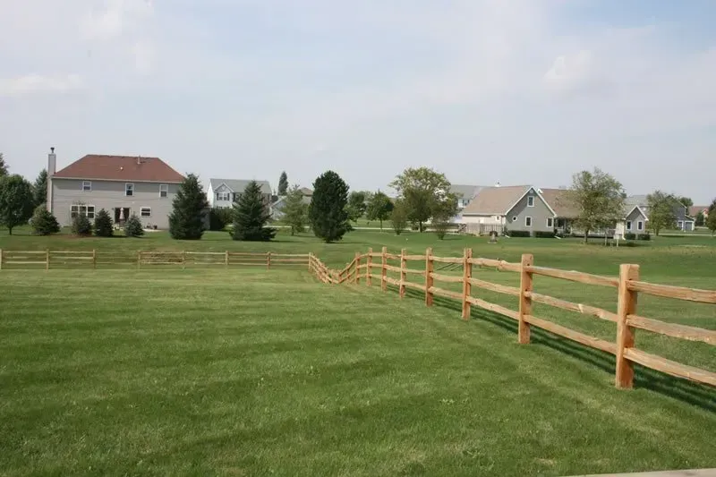 A wooden fence surrounds a lush green field with houses in the background