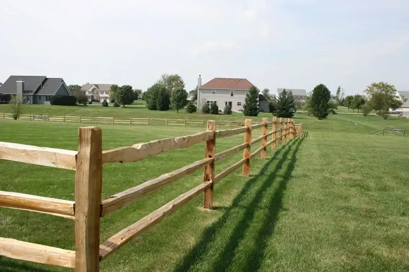 A wooden fence surrounds a lush green field with houses in the background.