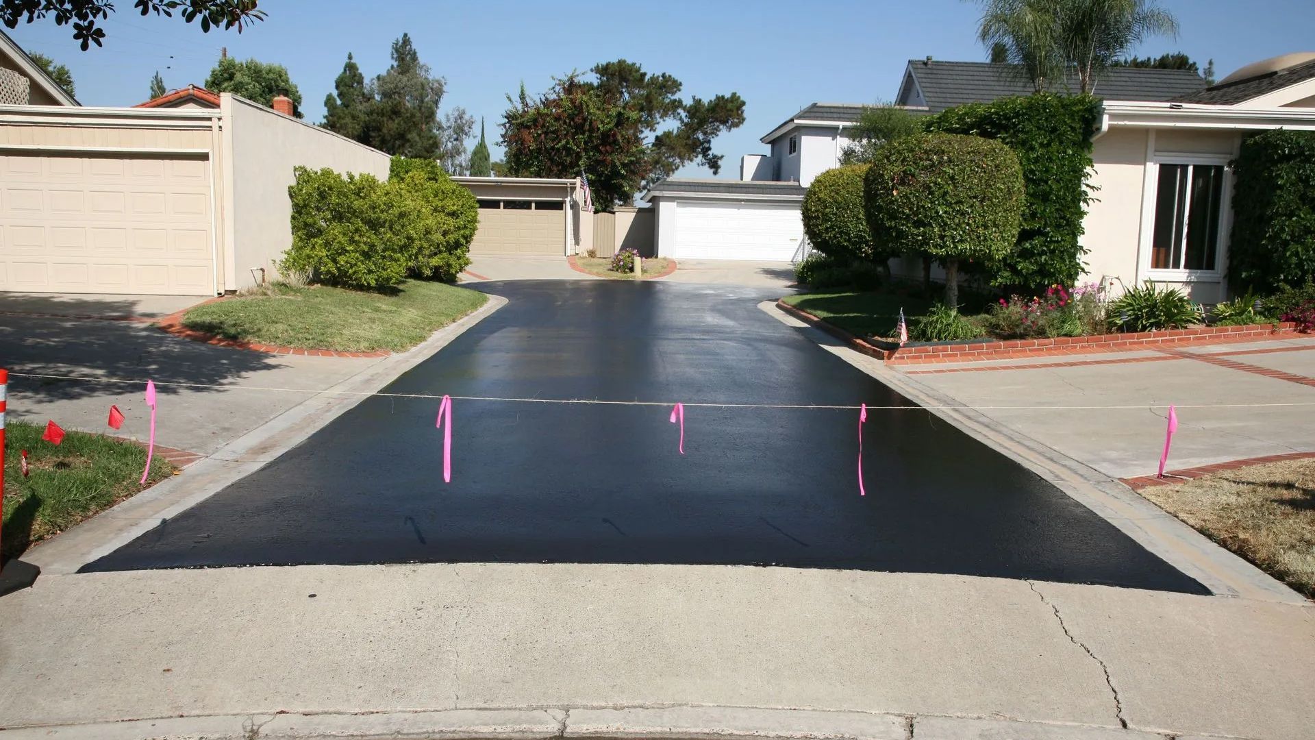 Freshly paved black driveway in a residential area, bordered by beige homes and pink survey flags.