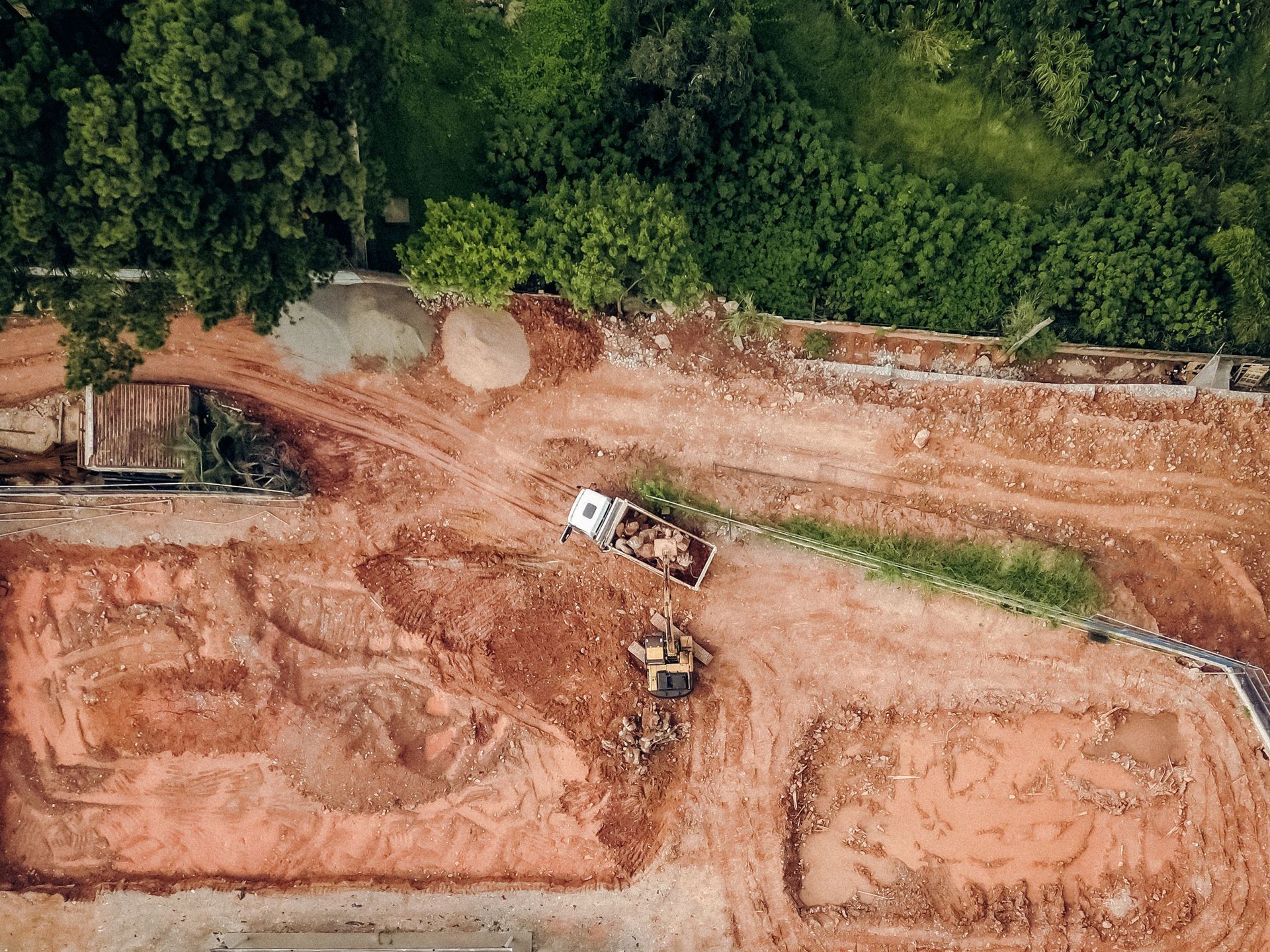 Aerial view of an earthworks construction site with excavators and dirt roads beside dense green trees