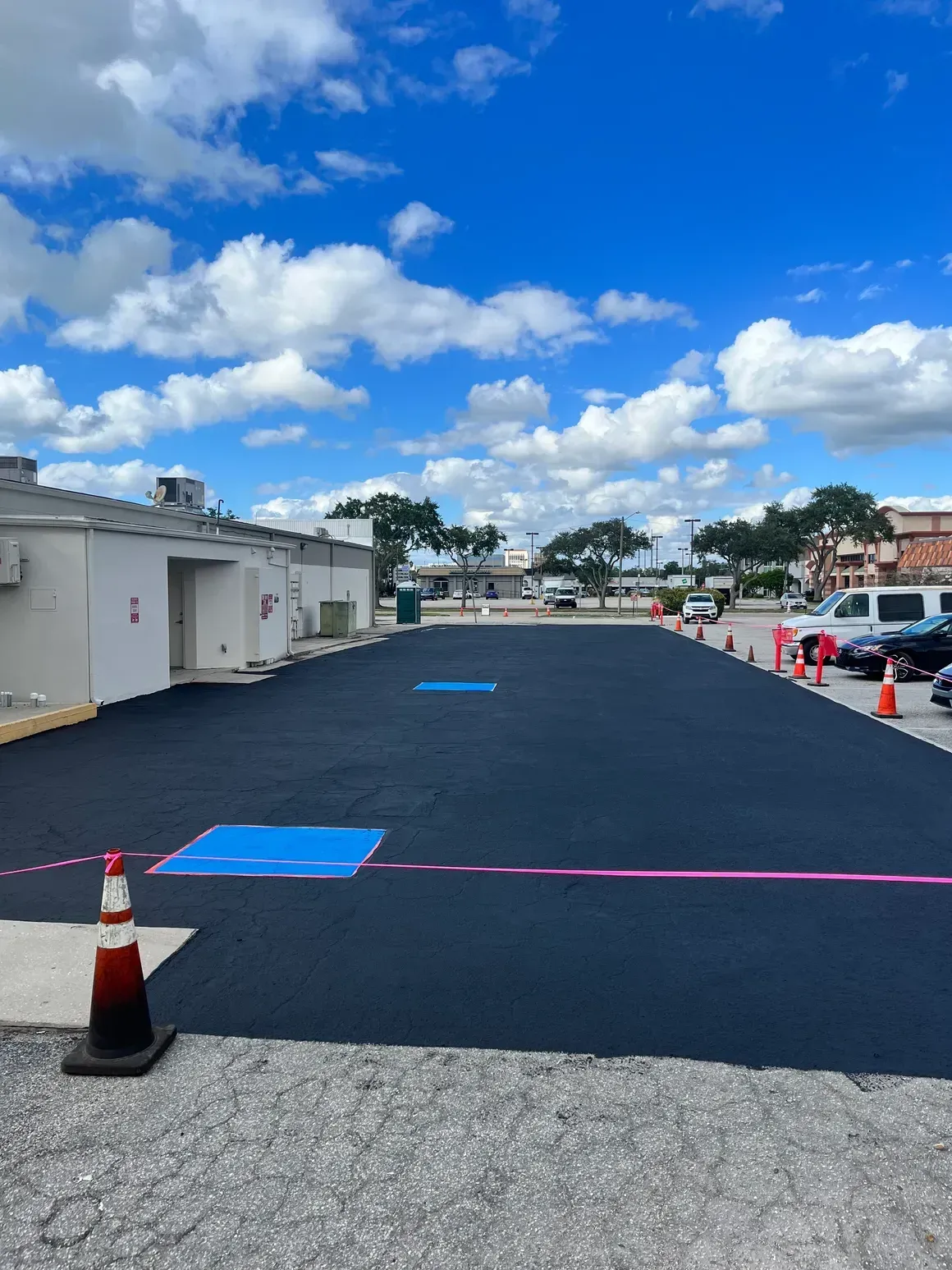 Empty parking lot with blue painted spaces, traffic cones, and buildings under a partly cloudy sky