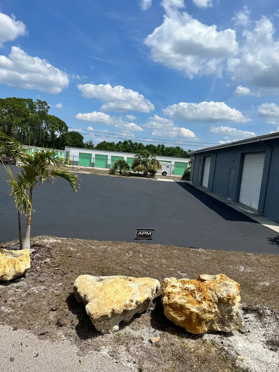 Empty gravel lot with rocks, a small tree, and a row of white storage units under a blue sky with clouds