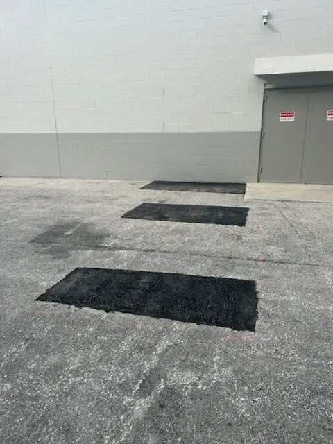 Paved lot beside a gray building with three black rectangular mats or patches on the ground