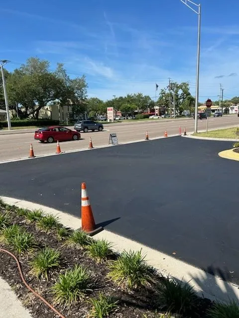 Freshly paved parking lot with orange cones, a red car, and trees under a clear blue sky