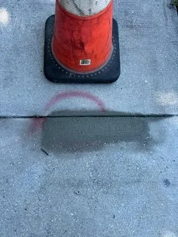 Orange traffic cone on concrete sidewalk beside a dark wet stain and crack line