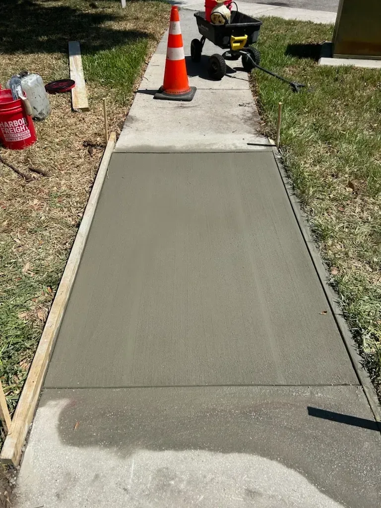 Freshly poured concrete sidewalk section with orange traffic cone and construction tools nearby