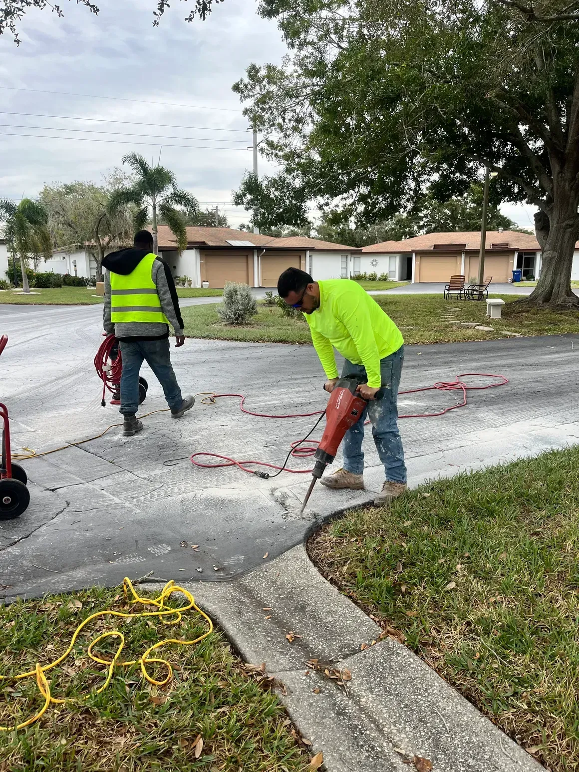 Workers in neon vests jackhammer cracked pavement on a residential street.