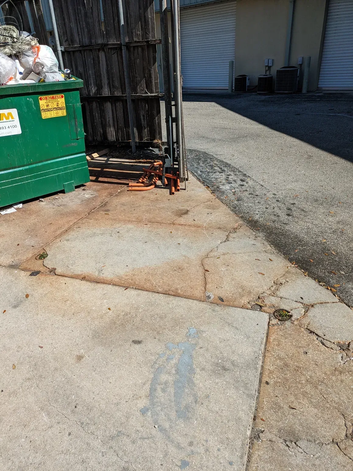 Cracked concrete beside a green dumpster and a chain-link gate in an alley
