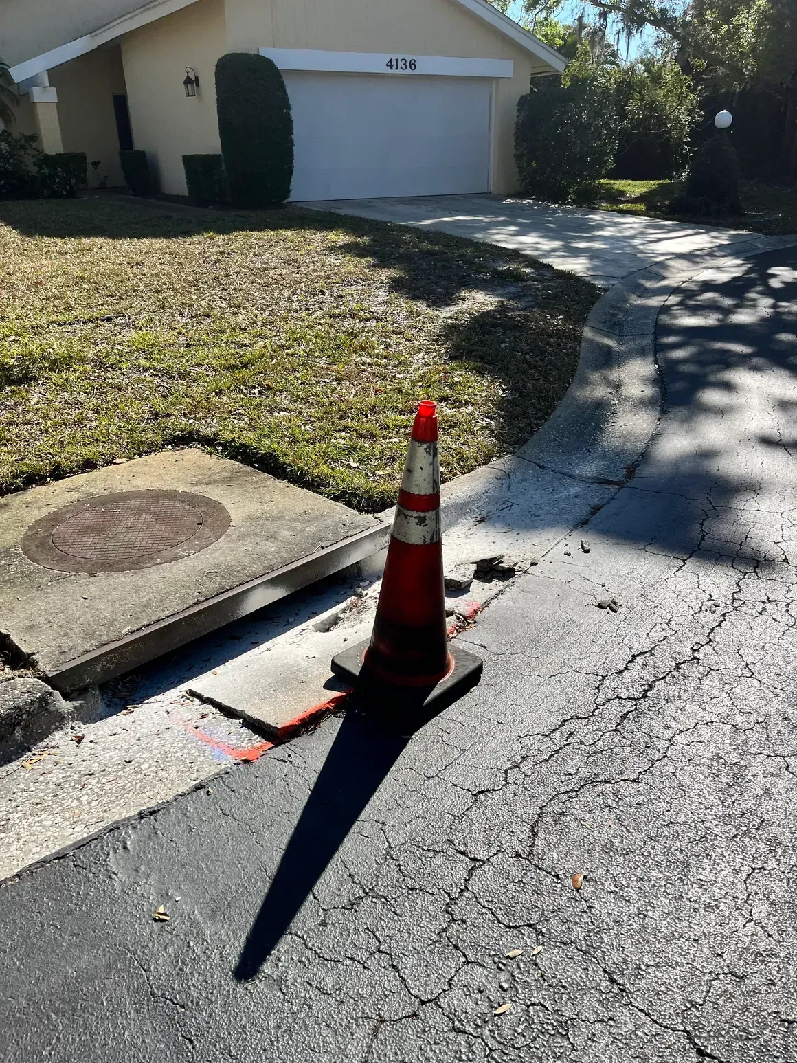 Orange traffic cone marking a road repair hole on a cracked residential street near a driveway
