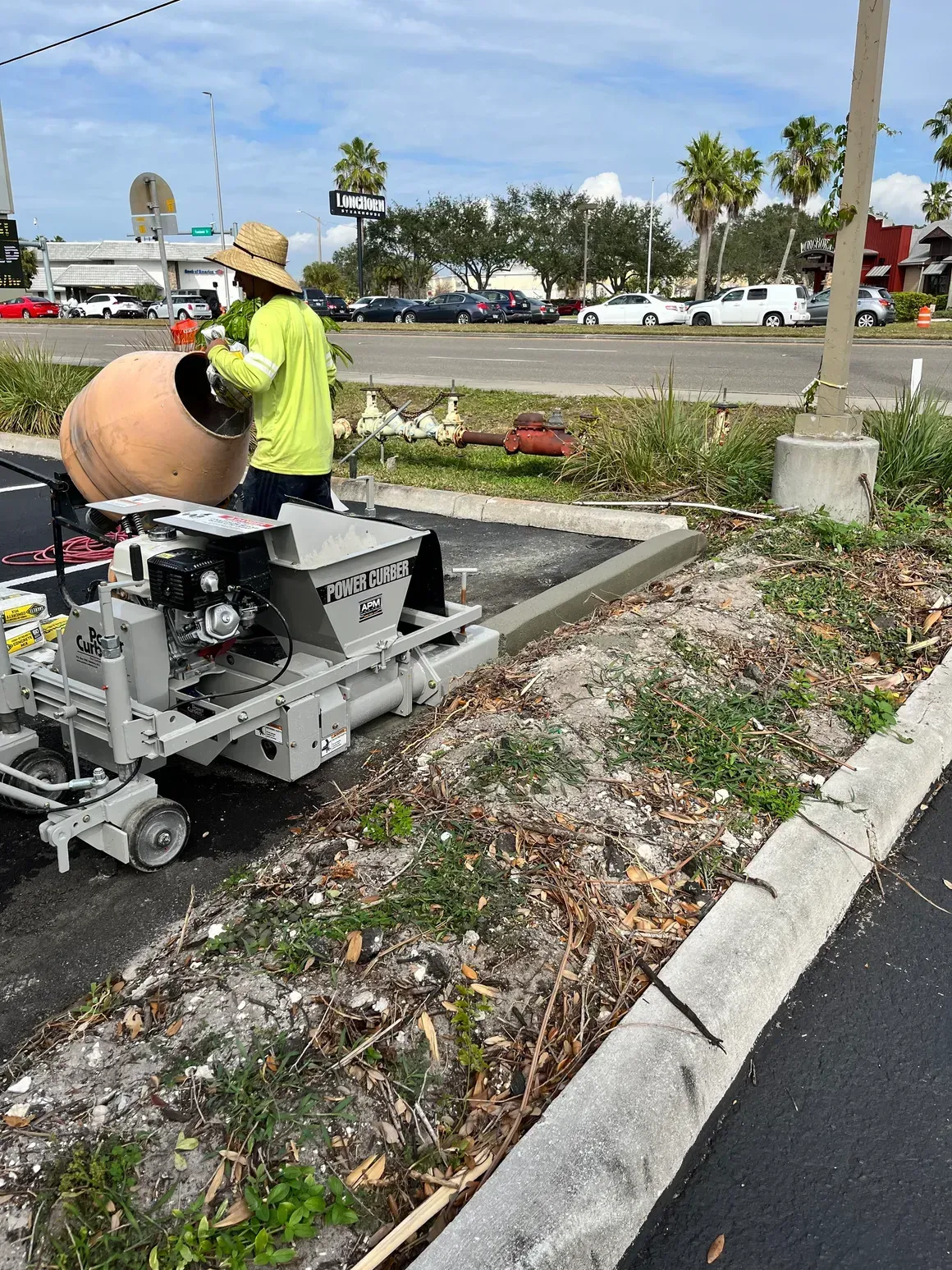 Worker in neon safety gear operates a curbside machine along a landscaped parking lot edge.