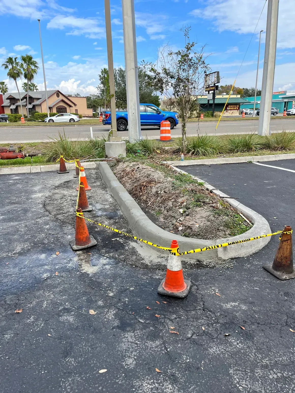 Damaged parking lot island roped off with orange cones and caution tape near a road.