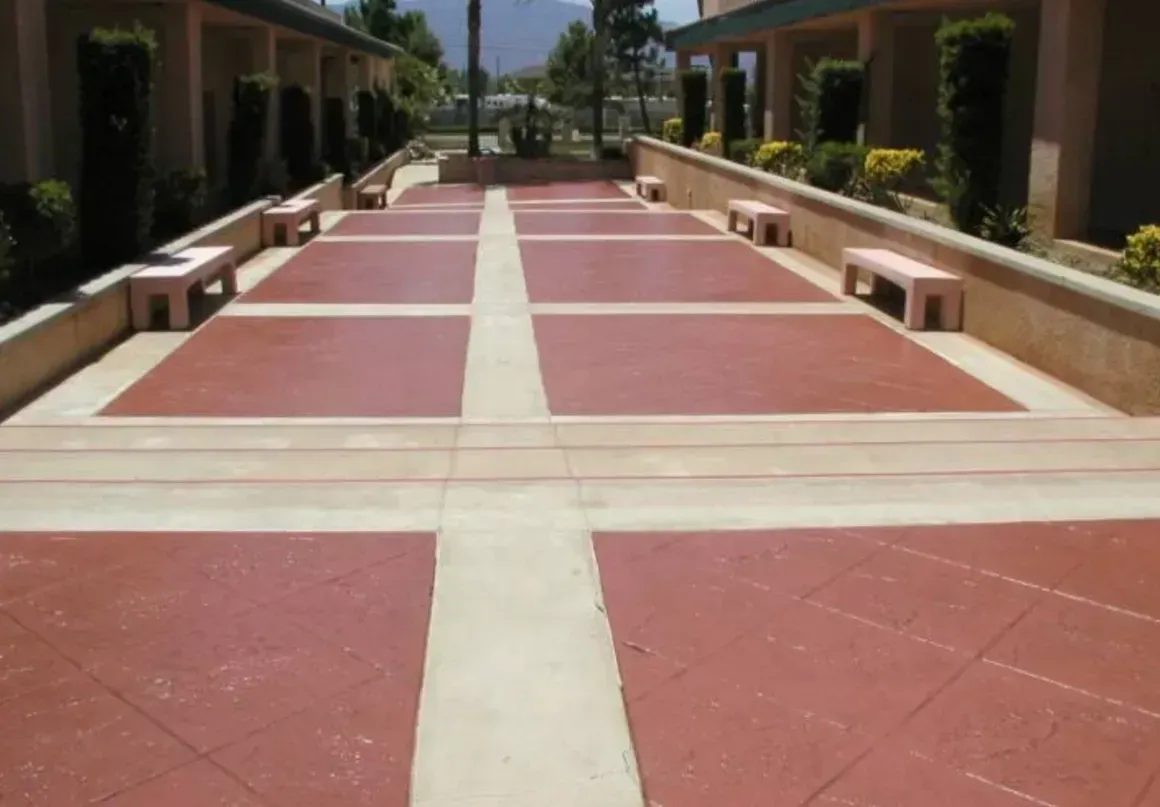 Red-tiled courtyard walkway with cream borders between low benches and buildings, leading toward a distant street