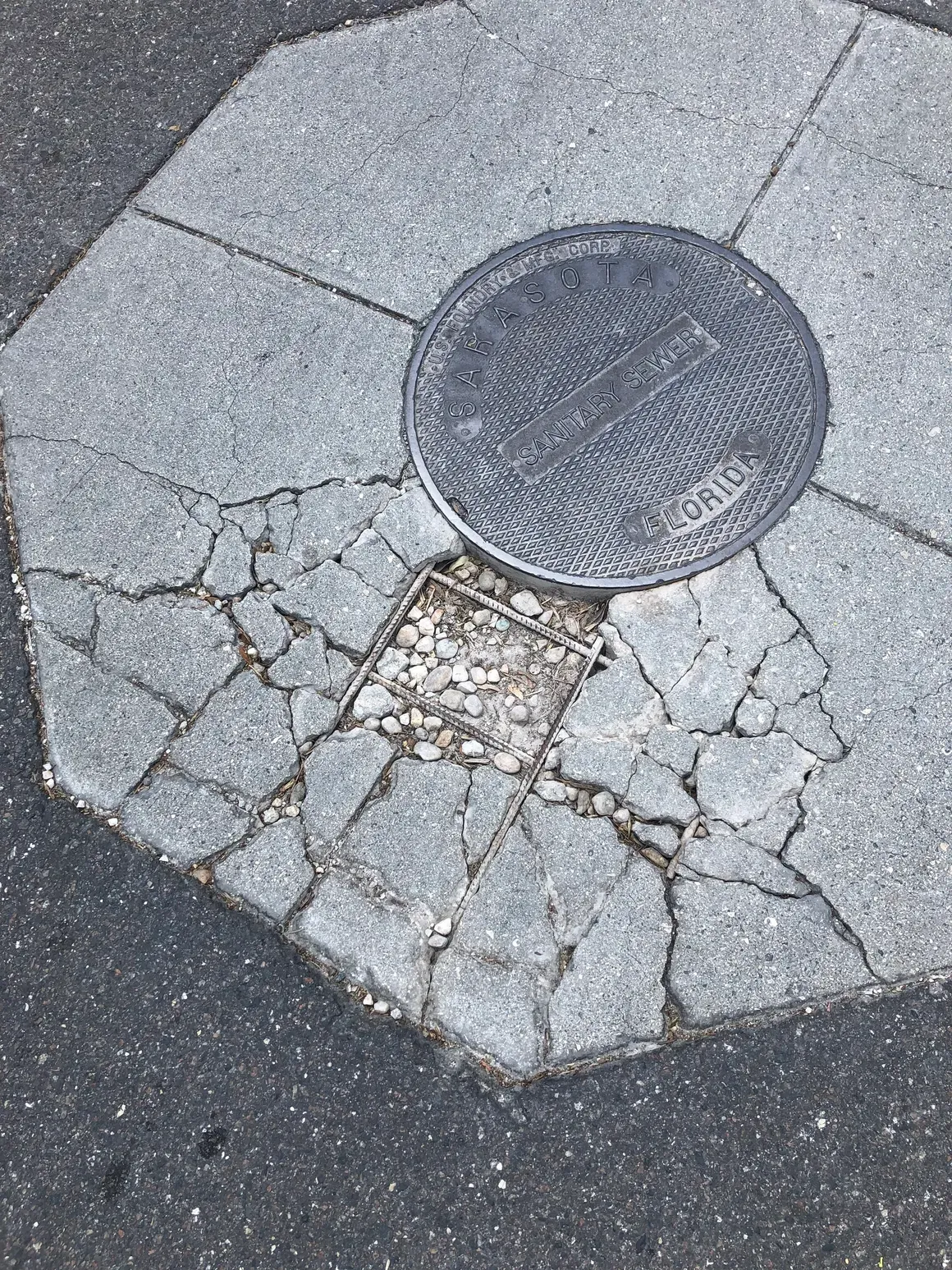 Damaged sidewalk with cracked concrete around a round manhole cover and exposed patchwork repair