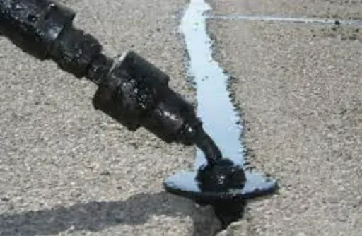 Close-up of a metal nozzle dispensing black sealant into a crack in pavement.