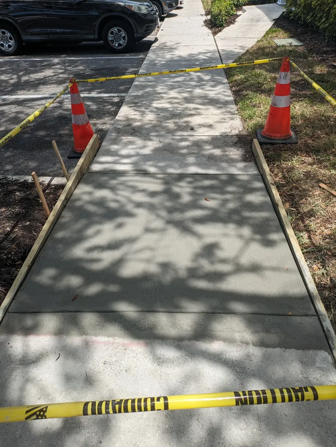 Sidewalk under repair, blocked by caution tape and orange cones near parked cars.