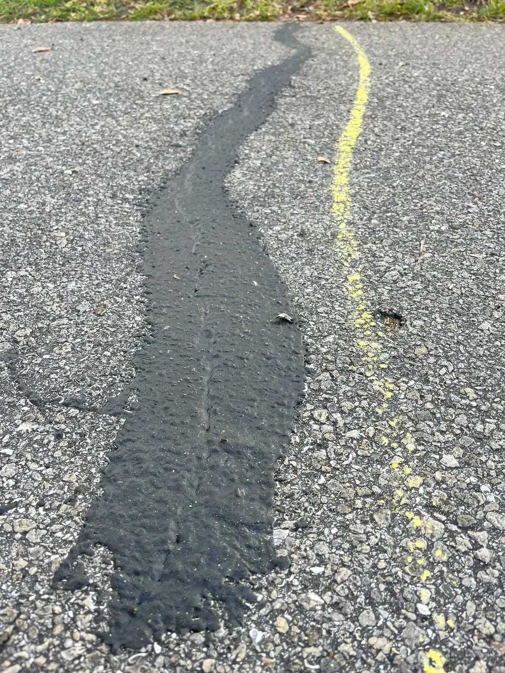 Cracked asphalt with a dark patch repair beside a yellow line on a gravel road surface