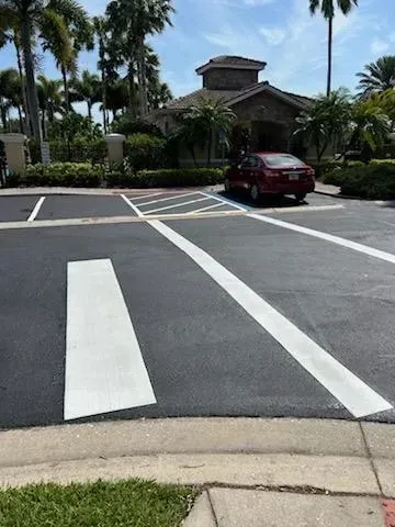 Parking lot crosswalk with white stripes leading to a red car near a building and palm trees.